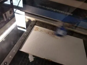 Sheet of white cardboard on a metal grate in a laser cutter behind dirty glass. There is a blur of a moving laser cutter head over the center third of the cardboard. A completed etching of the Dominion logo can be seen as a pale brown on the cardboard's surface.