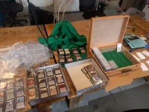 Resting atop a wooden table which fills the frame, from left to right there is a stack of two sheets of card sleeves full of Dominon cards, the bottom half of a Dominion box full of card sheets which are them selves full of Dominion cards, the top half of a Dominion box with three stacks of dominion cards, a light wooden box which is open, with a green felt bottom split into six columns by strips of clear acrylic, one of the columns is full of dominion cards with a second one about half full, and finally five stacks of white card paper which are barely in frame. On the right side of the table there is also a jumble of empty card sleeves.