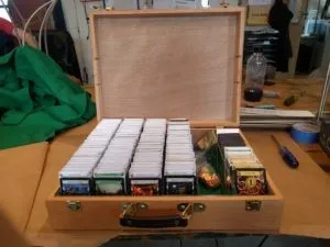 A light wooden box sits dead center, filling the bottom half of the frame. Its open lid fills the top half. The box is filled with dominion cards arranged in four of the six columns and separated by white card stock with card titles. The second to left column contains little plastic bags of glittering bronze tokens, and the last column is about half full of cards separated in the same manner as the first four.
