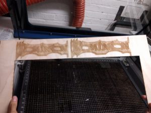 Strip of birch plywood with two blury laser etched dominion logos filling it's length runs across the center of the picture. Behind it is the metal latice of a laser cutter.