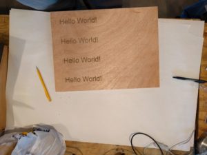 Wooden rectangle sites atop white paper. The words "Hello World!" are laser etched into four rows filling its left half.