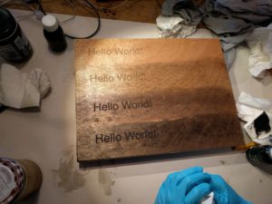 Wooden box with the words "Hello world" etched in four rown down the left side, sits on white paper with a bright light shining down on it. The wood has been stained in four rows with increasingly dark wood stains / waxes going from a golden honey colour to a brushed black hue.