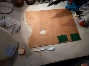 A rectangular wooden pallet has two small squares of green felt glued to the bottom right corner of the palette. A jug of wood glue, can of spray adhesive, and hand holding a hot glue gun are just out of frame.