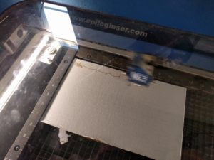 Sheet of white cardboard on a metal grate in a laser cutter behind dirty glass. There is a blur of a moving laser cutter head over the center third of the cardboard. The beginning of an etching of the Dominion logo can be seen as a pale brown on the cardboard's surface.