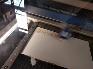 Sheet of white cardboard on a metal grate in a laser cutter behind dirty glass. There is a blur of a moving laser cutter head over the center third of the cardboard. A completed etching of the Dominion logo can be seen as a pale brown on the cardboard's surface.