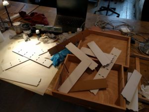Seen from above, the right half of a wooden table is covered in two disjoint halves of a wooden box, the left is covered in white paper. Strewn over everything is a set of cardboard inserts which are white on one face and brown on the other.