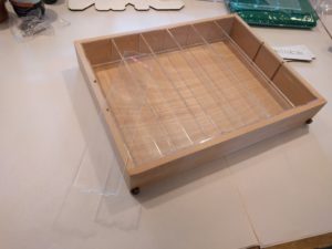 Light wooden box sits atop white paper at a slight angle filling the upper two thirds of the frame. The box is divided into six columns by strips of clear acrylic which are the same height as the box itself. An additional strip of clear acrylic rests atop the left edge of the box hanging prosperously over the side.