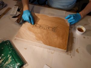 Light brown wooden box sits in middle frame at a slight angle atop white paper. It is mostly covered in a honey brown wax. The Dominion logo is etched in its center. Two hands connected to arms which lead off the top of the picture, are wearing blue nitirle gloves and rubbing additional wax into the surface.