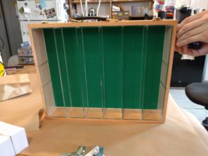 Light wooden box with a green felt base rests on its edge, filling the frame. The box's inside is divided into six vertical columns by clear acrylic. Two hands in the upper right of the frame can barely be seen screwing something into its side.