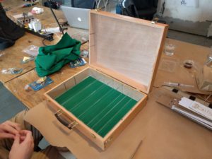 Open light brown wooden box resting on butcher paper on a wooden table. The bottom of the box is green felt and it is divided into six columns by clear acrylic strips.