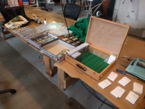 A wooden table runs at a sharp angle from the top left to the bottom right of the frame. From left to right there are: sheets of card sleeves full of dominion cards, the bottom half of a dominion box half full of full card sheets, the top half of a dominion box with three stacks of dominion cards in it, a light wooden box which is open, its base green felt, divided into six columns by clear acrylic the left most of which is full of dominion cards and dividers, finally six stacks of white card stock with dominion rules text printed on them.