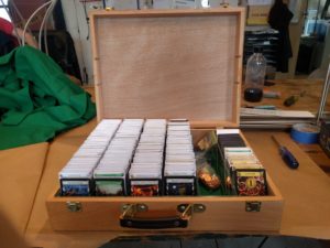 A light wooden box sits dead center, filling the bottom half of the frame. Its open lid fills the top half. The box is filled with dominion cards arranged in four of the six columns and separated by white card stock with card titles. The second to left column contains little plastic bags of glittering bronze tokens, and the last column is about half full of cards separated in the same manner as the first four.